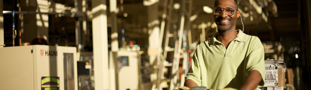 Smiling black manufacturing worker with protective eye wear and a sage green polo shirt with machinery in the background