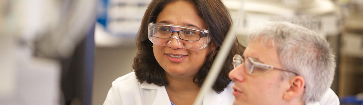 Female scientist with brown shoulder-length hair and a male scientist with grey hair both in protective glasses and white lab coats looking at a screen in a lab