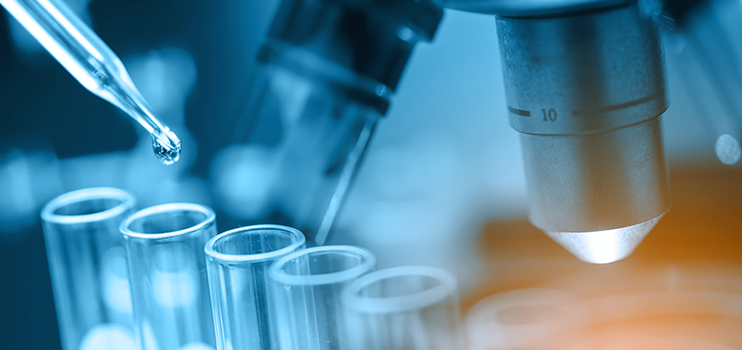 Close up of a microscope with a rack of six test tubes in front and a dropper poised over the second vial in the rack.  Entire image is shaded in blue light.