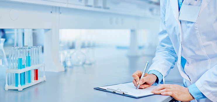 White male scientist in blue shirt and white lab coat making report notes in a lab with test tubes in a rack on the counter holding translucent blue and red fluid.