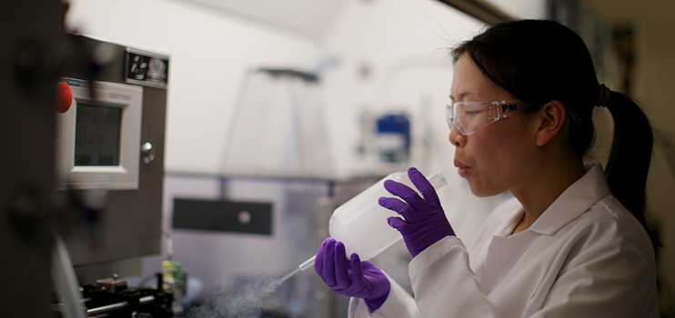 Asian female scientist in white lab coat and purple gloves blowing through one opening of an oblong glass flask with smoke exiting the other side