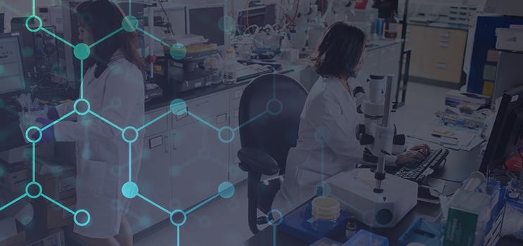 Asian female scientist in white lab coat seated in black chair before a computer and typing on a grey keyboard in a lab with second female scientist standing behind her at a countertop carrying a container with vials to a computer station.