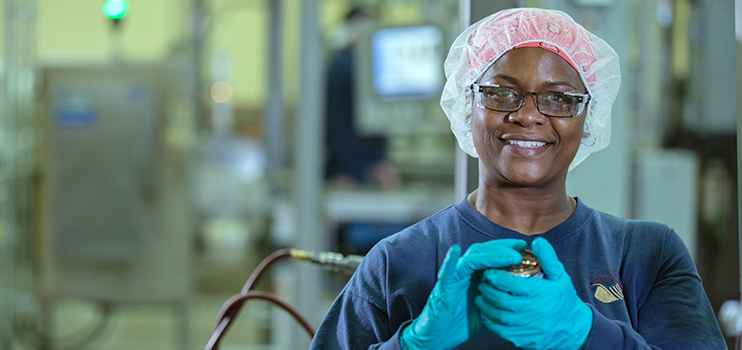 Smiling black female factory worker in dark-rimmed glasses with a patterned red head wrap covered by a transparent white bonnet wearing a navy sweatshirt and blue gloves. Background is blurred.
