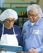 Two factory workers side by side in white hair nets with the person on the left in a white apron over a navy blue long-sleeved top and the man on the right in the blue long-sleeved buttoned shirt. They are reviewing a document held by the aproned worker.