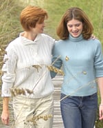 White mother in a white long-sleeved sweater walking outside with her daughter who's wearing a light blue turtlenecked sweater and with jeans.