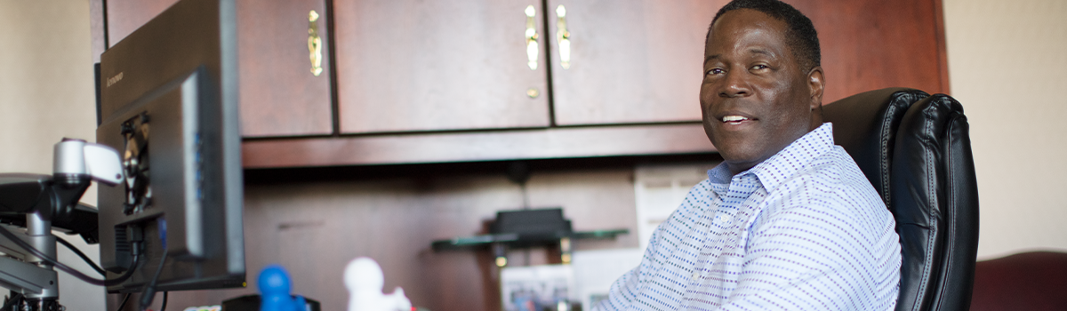 Smiling black man sitting in an office with wood cabinets, on a black highbacked padded office chair, wearing a long-sleeved, collared buttoned-down light blue shirt with horizontal rows of dots in blue and purple.
