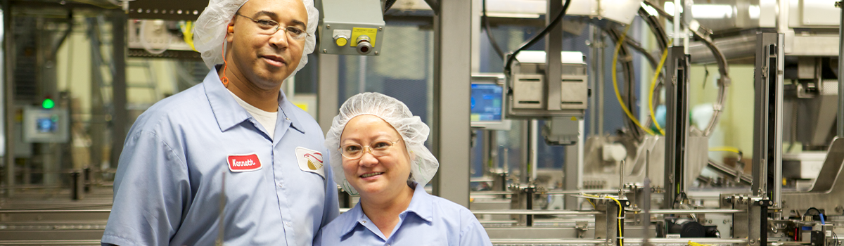 Two factory workers, both in white hair nets and blue long-sleeved buttoned-down shirt standing in front of machinery.