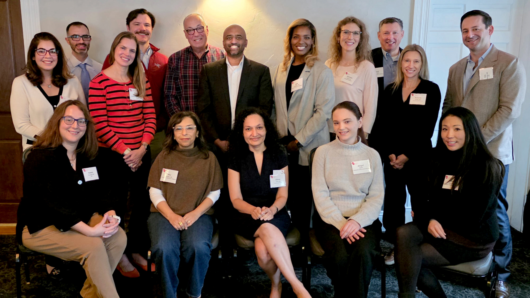 Group photo of approximately 18 smiling adults wearing name badges, posed in rows indoors.