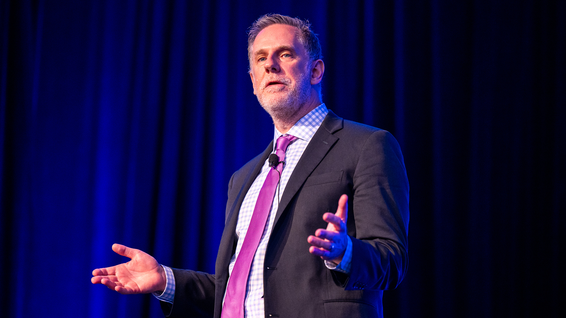 Conference speaker in a dark suit and pink tie stands onstage under blue lighting, gesturing while addressing an audience.