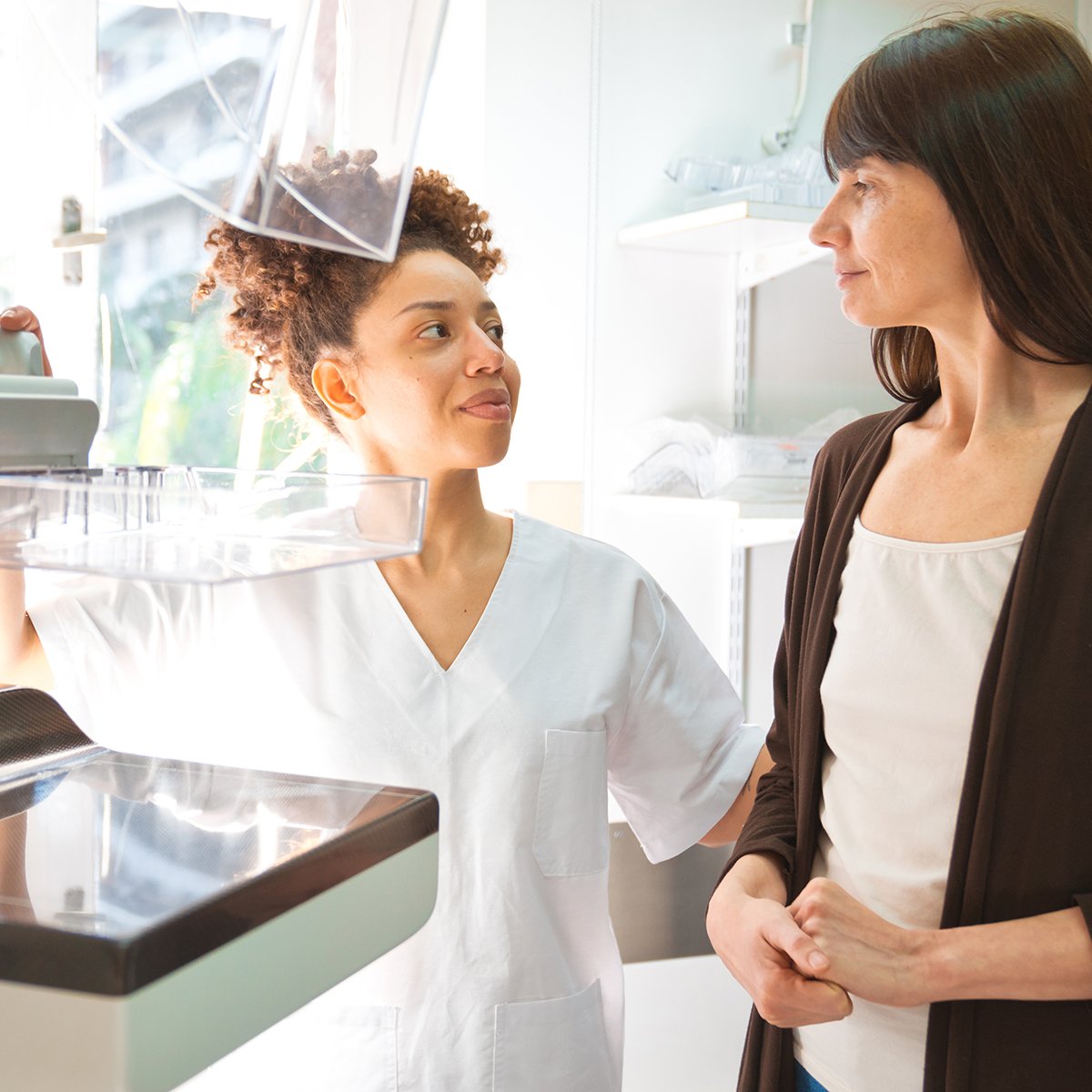 African-American radiologic technologist with female patient standing in front of mammography equipment prior to performing a mammogram.
