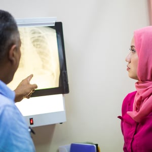 A woman in a pink hijab looks at a chest x-ray as a Black radiologist points to something on the scan. 