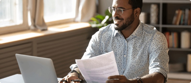 A person sitting behind their desk while looking at their laptop and holding a document.