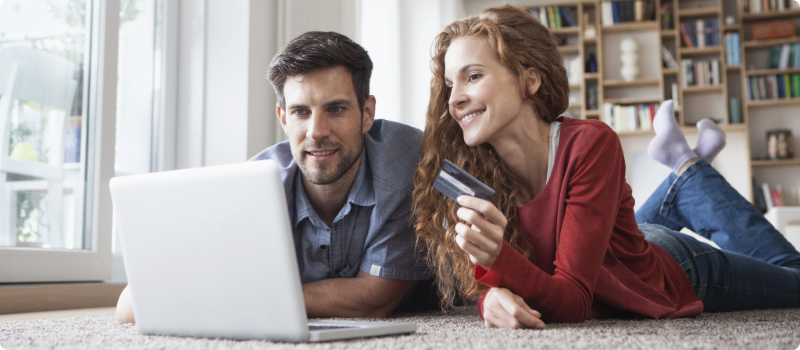 A couple lying on their floor together in front of a laptop while holding a credit card. 