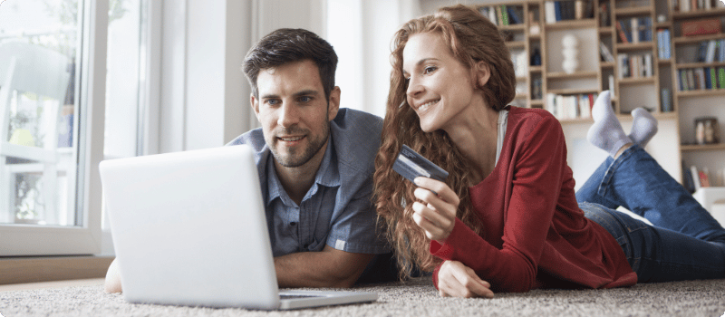 A couple lying on their floor together in front of a laptop while holding a credit card.
