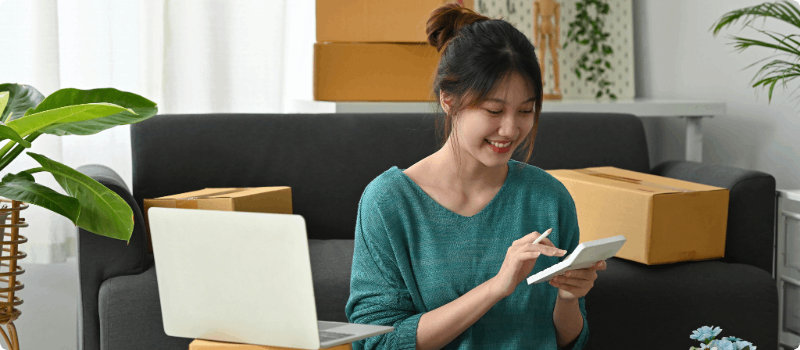 Person in her living room using a calculator in front of a laptop while surrounded by boxes.