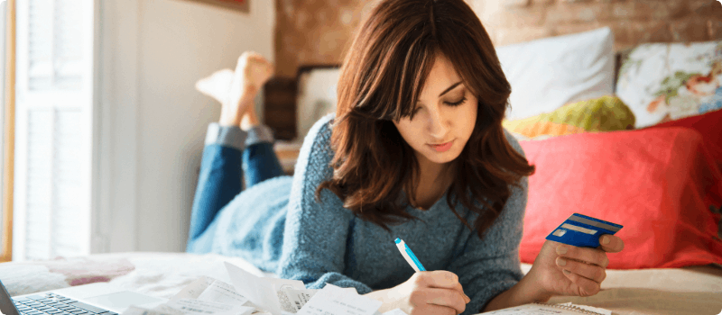 Person holding a credit card and writing in a notebook while lying on her bed.