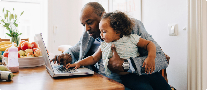 A father working on his laptop at home while holding his young child.