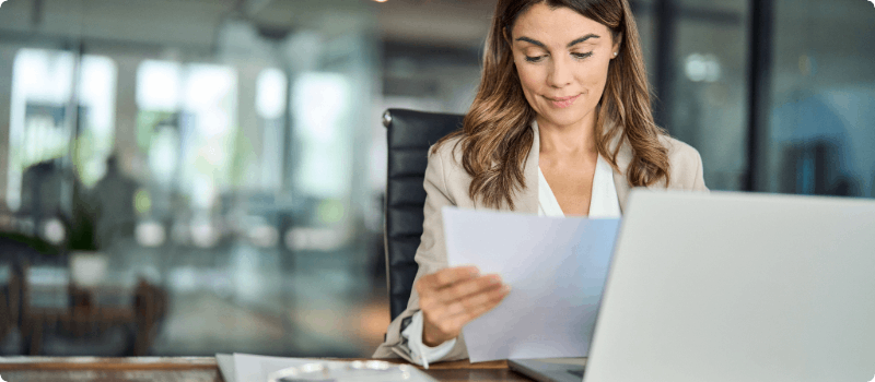 A person sitting at her desk in front of her open laptop while holding a document.