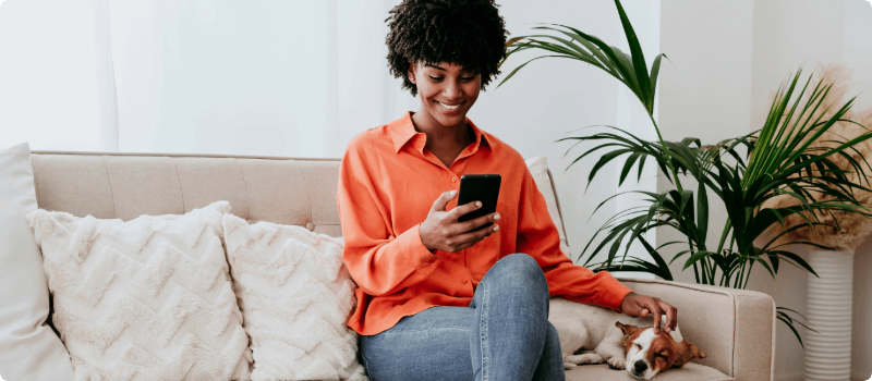 A happy person, sitting on her couch and looking at her phone while petting her dog. 