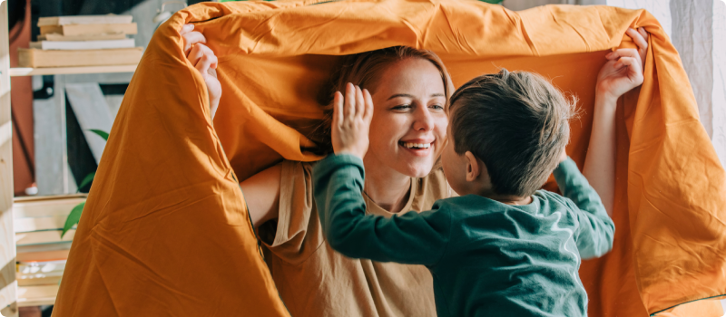 Playful mother holding blanket playing with son on bed at home.