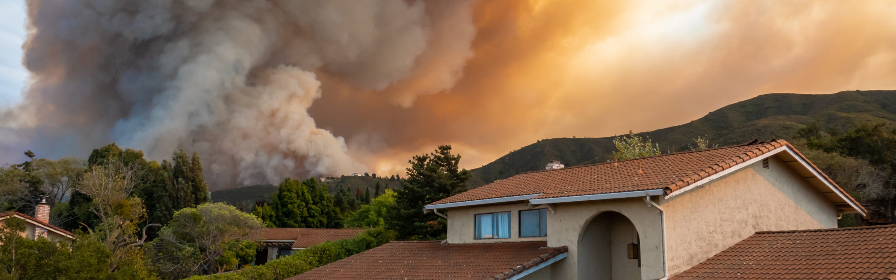 A home on a hillside with a wildfire growing in the background.