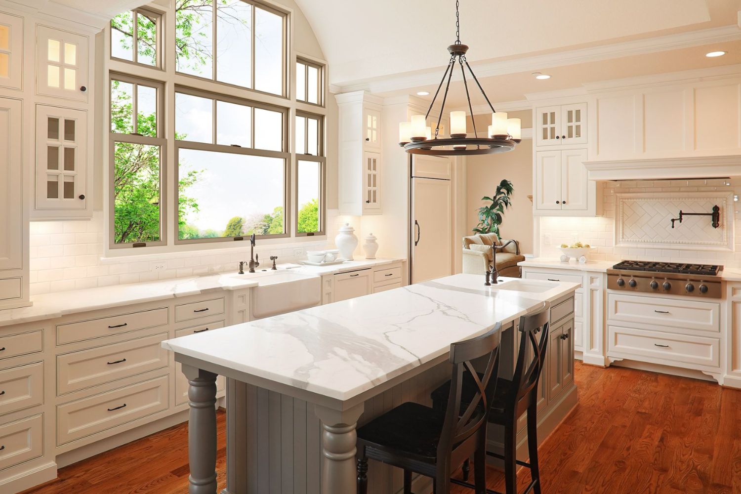 Brightly lit kitchen with wood floors, a marble-top island in the middle of the room, surrounded by clean, white cabinetry. There is an arrangement of windows letting in light behind the kitchen sink.