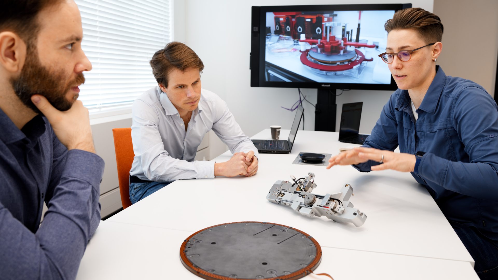 Three ASML employees examine a piece of machinery in a meeting room, a computer screen in the background. 