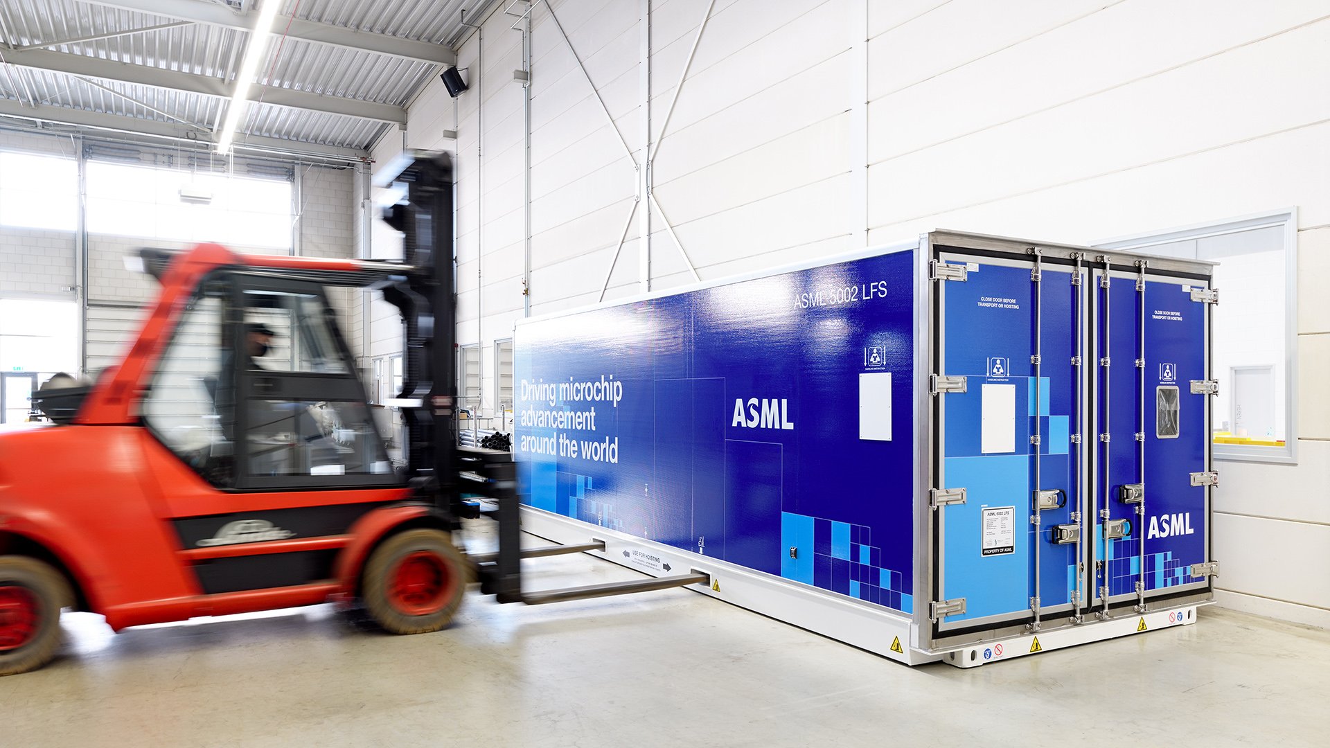Inside a warehouse, a red forklift moves towards an ASML shipping container.