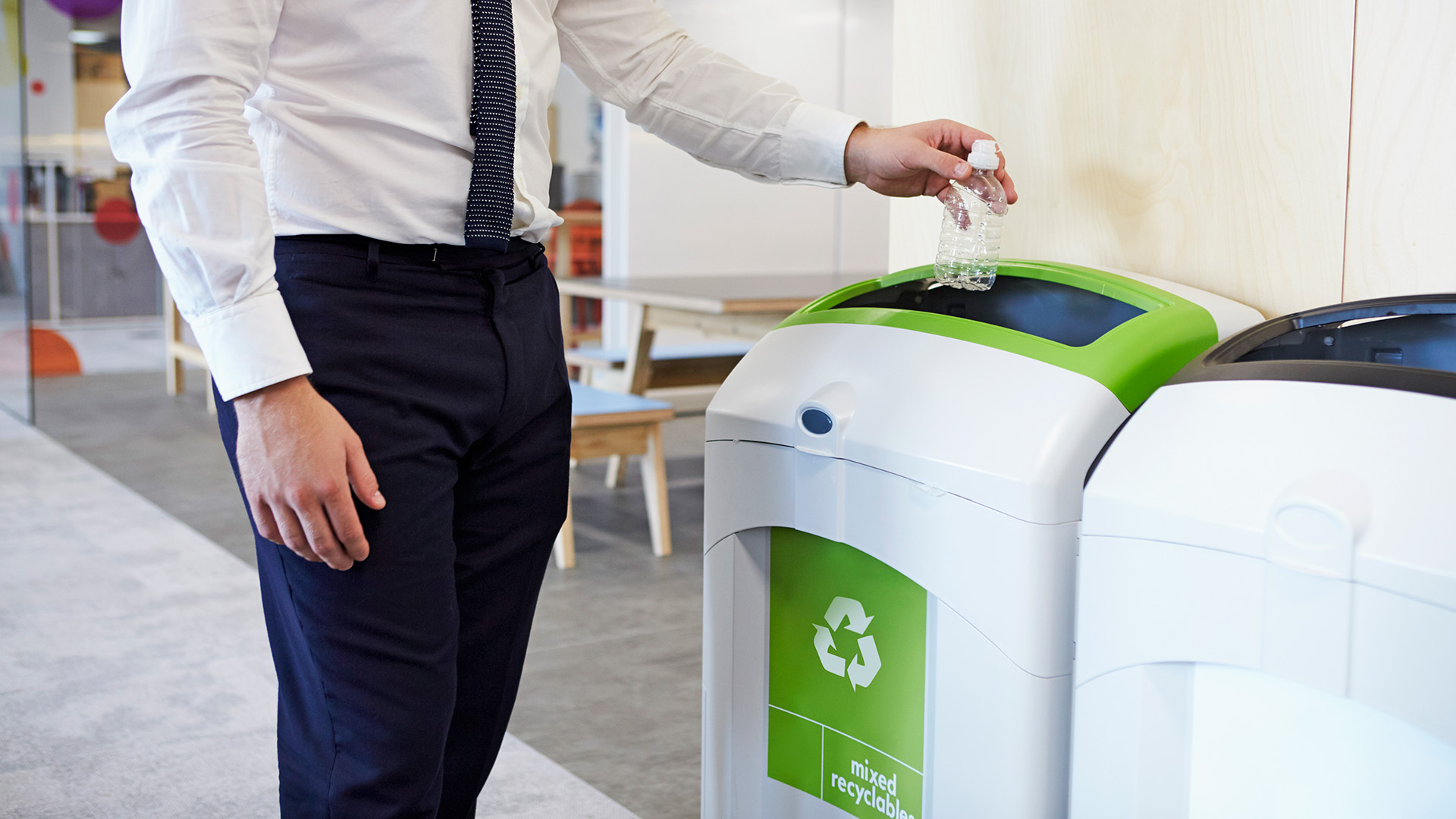 Partial view of an individual in business attire recycling waste into a green bin.