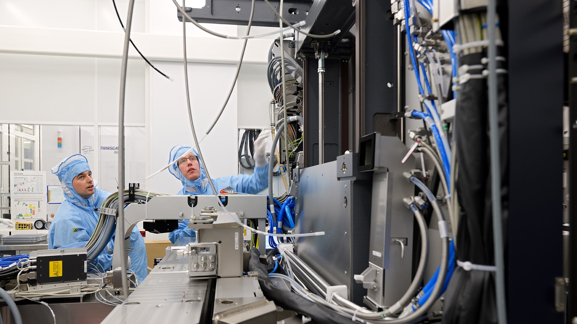 Two ASML technicians wearing blue cleanroom suits examine a lithography machine.