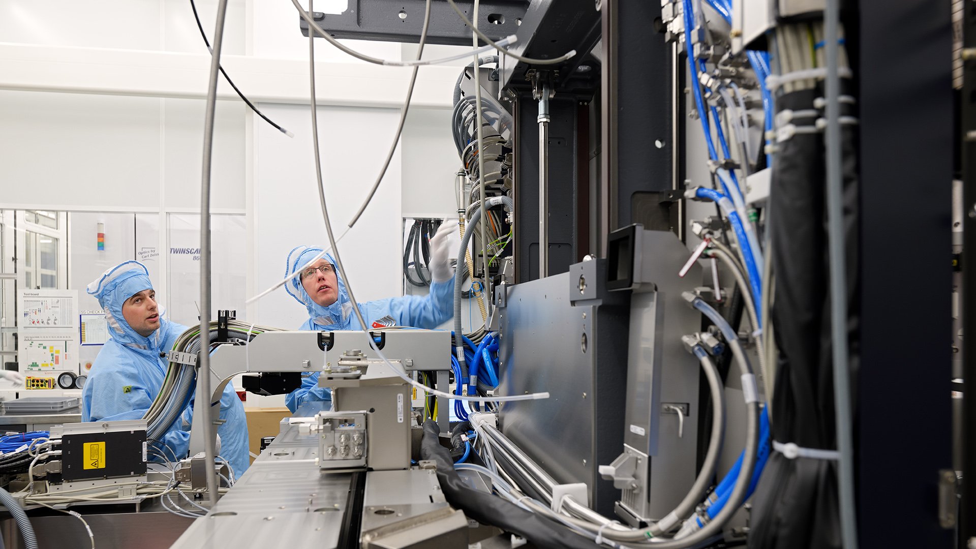 Two ASML technicians wearing blue cleanroom suits examine a lithography machine. Two ASML technicians wearing blue cleanroom suits examine a lithography machine.