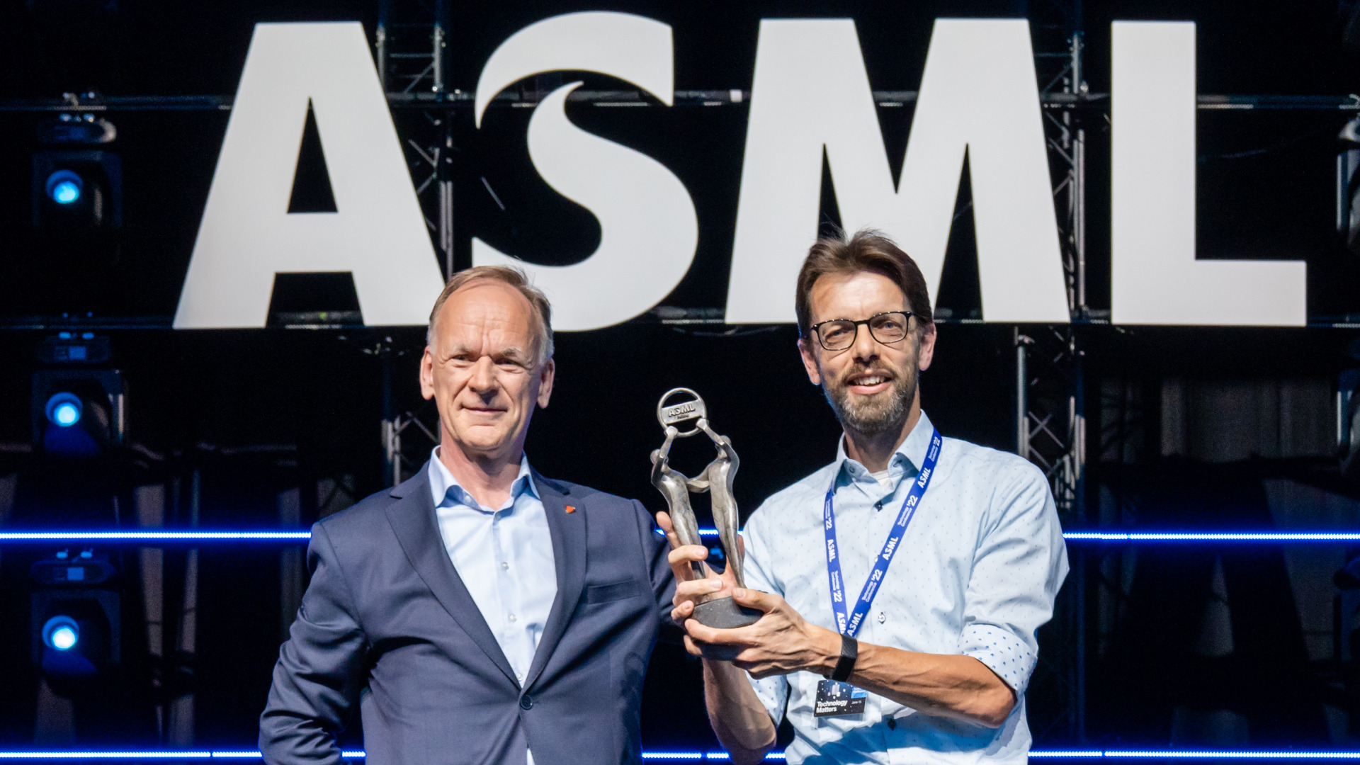 ASML Fellow Antoine Kempen stands to the right of senior vice president for technology Jos Benschop. He holds up his fellow award. They both stand under the ASML logo.