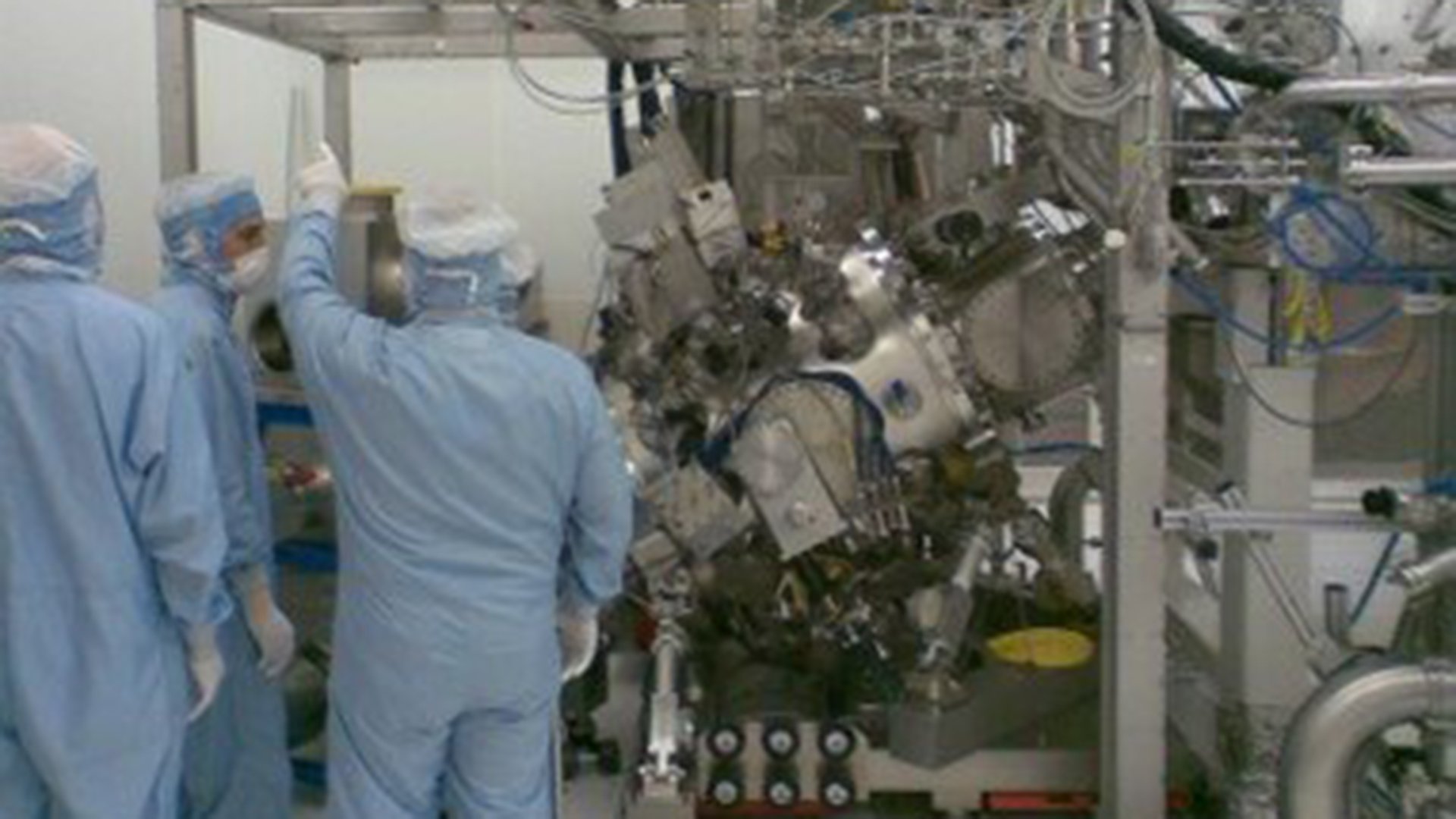 Three ASML cleanroom engineers stand next to a Cymer EUV light source. Three ASML cleanroom engineers stand next to a Cymer EUV light source.