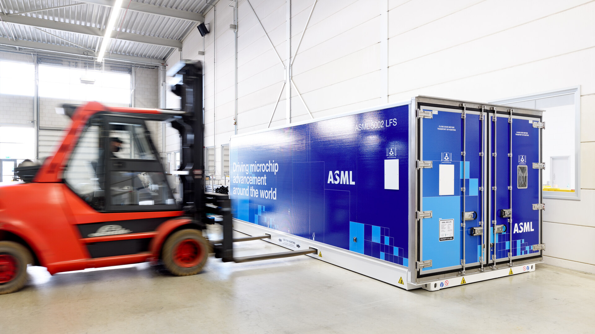 A forklift inside a warehouse approaches a large blue container bearing the words “ASML” and “Driving microchip advancement around the world”.