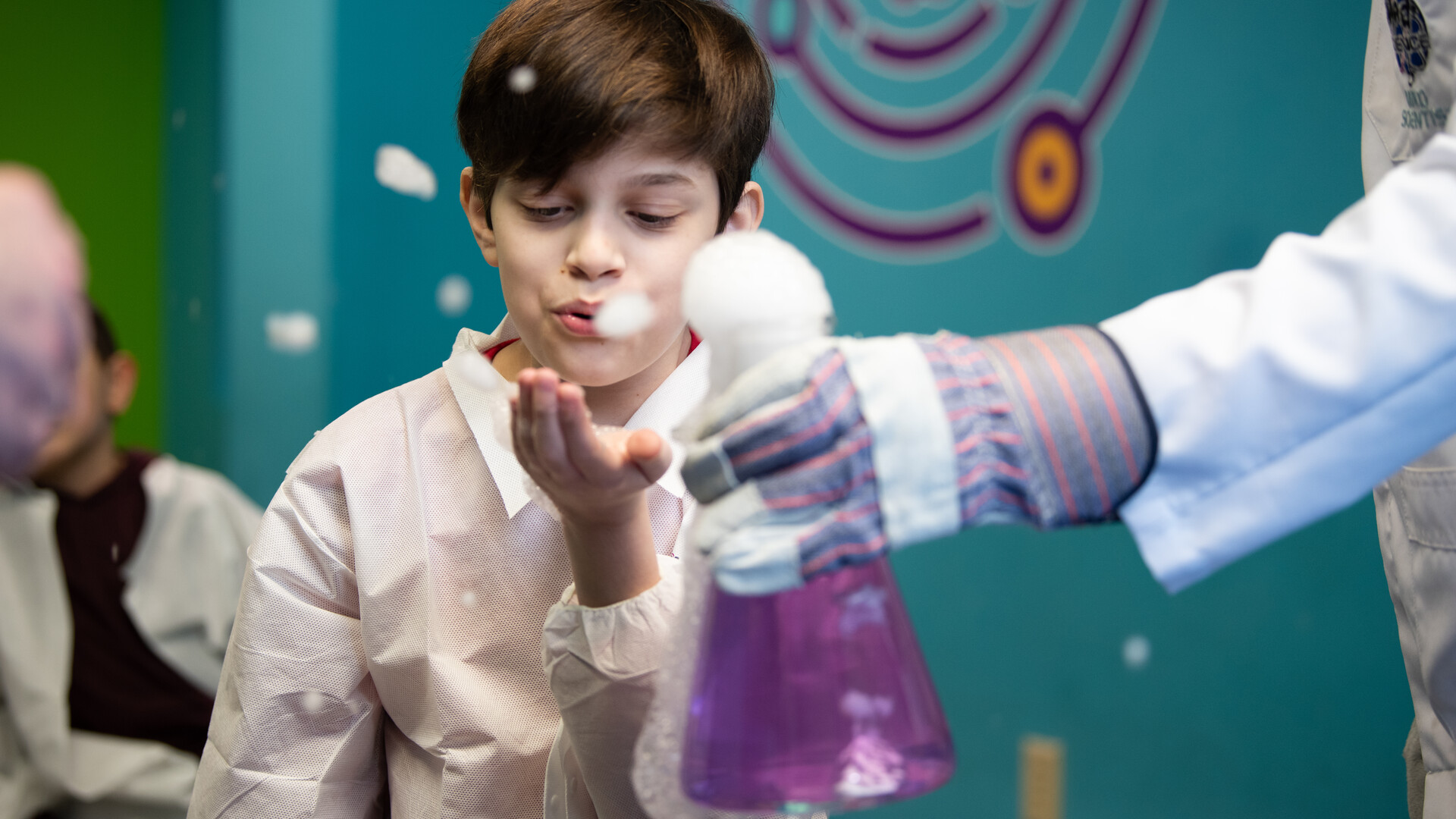 A child is seen blowing at the foam in his hand as a result of an overflowing flask full of bubbly purple liquid during a science experiment.