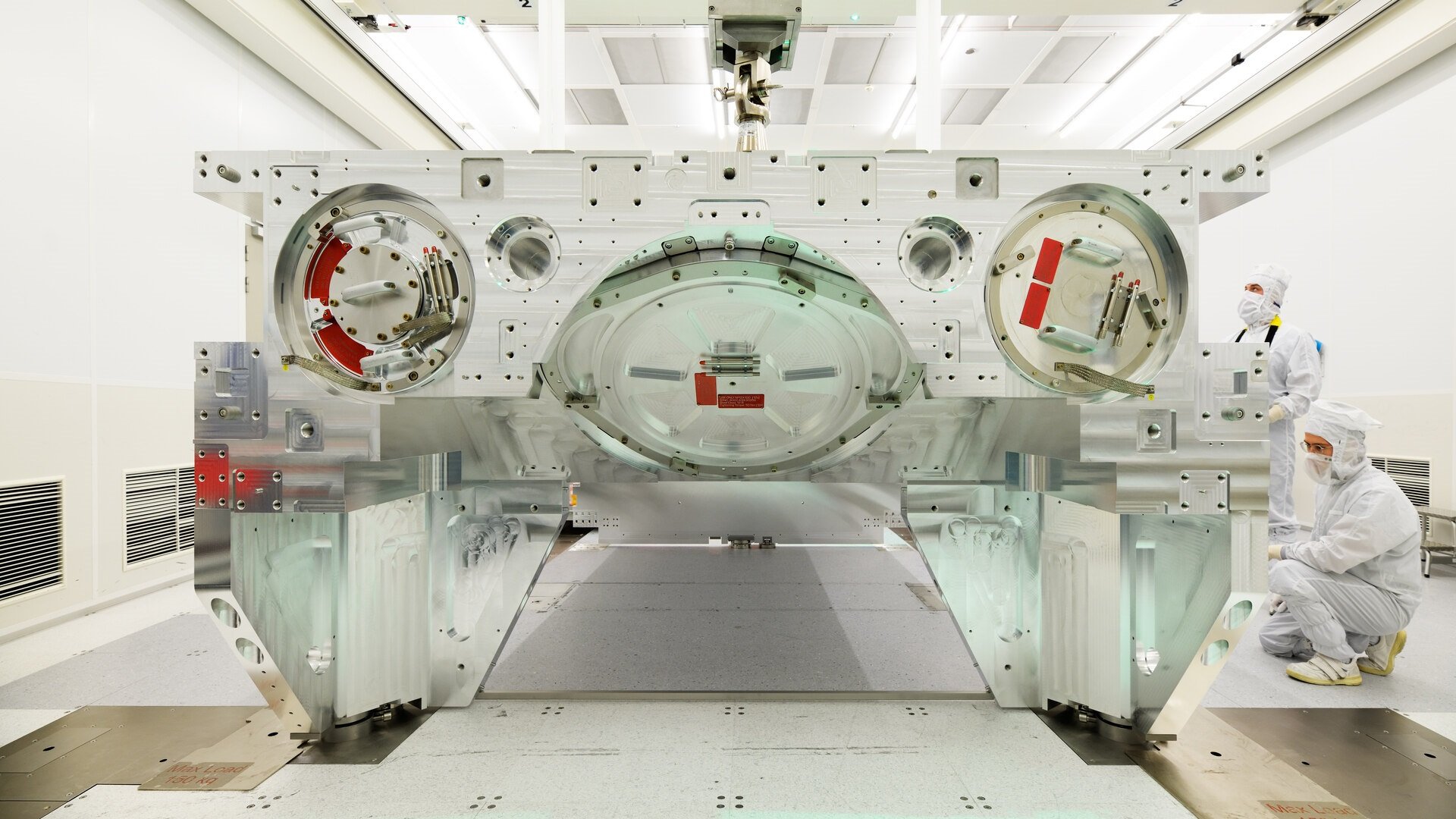 Two people in cleanroom suits observe while a crane hoists a bottom module frame Two people in cleanroom suits observe while a crane hoists a bottom module frame