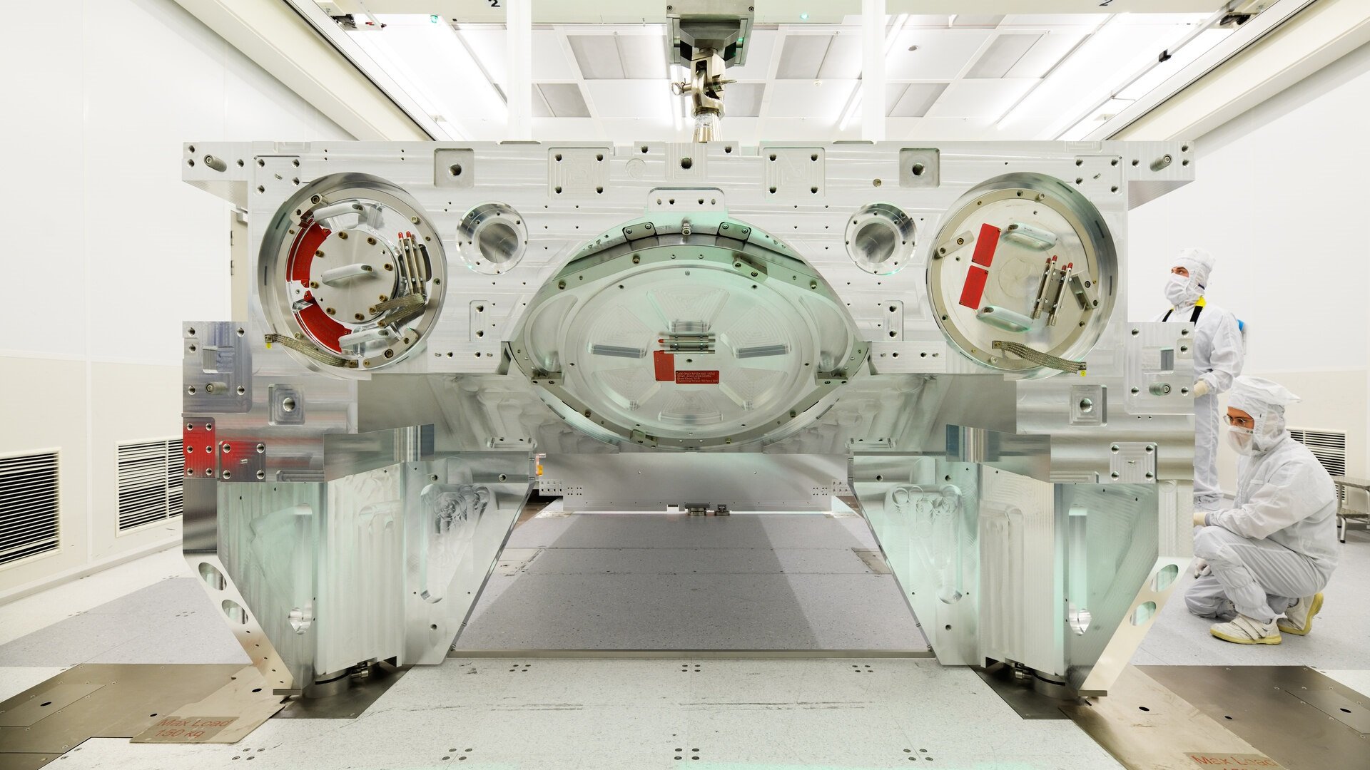 Two people in cleanroom suits observe while a crane hoists a bottom module frame