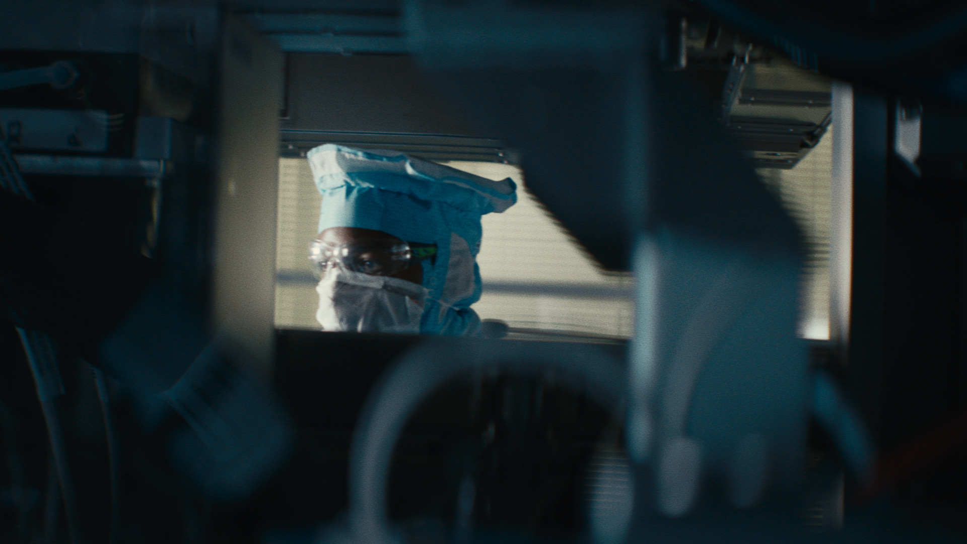 The camera looks through a machine to see the face of a man wearing a cleanroom suit, a mask and safety goggles.