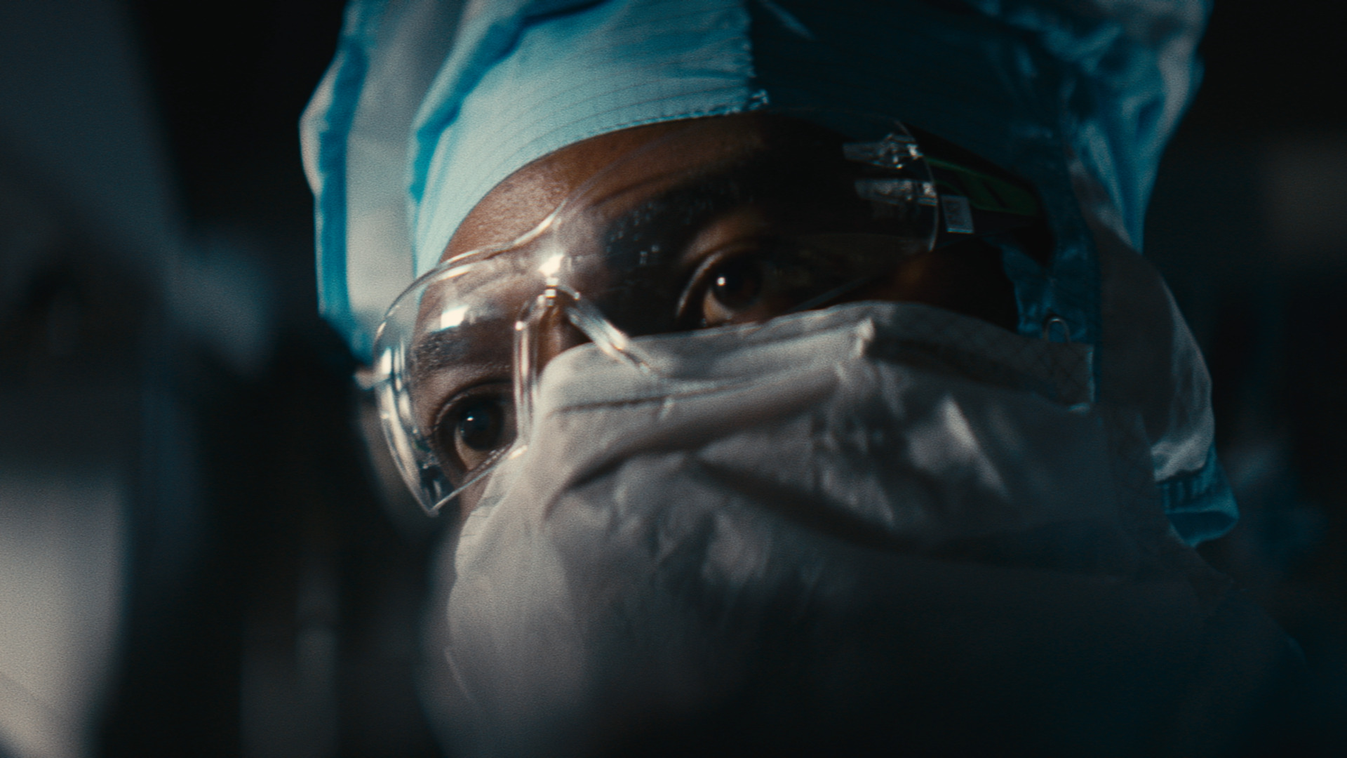 The face of a man wearing a cleanroom suit, mask and safety goggles looking into a machine.