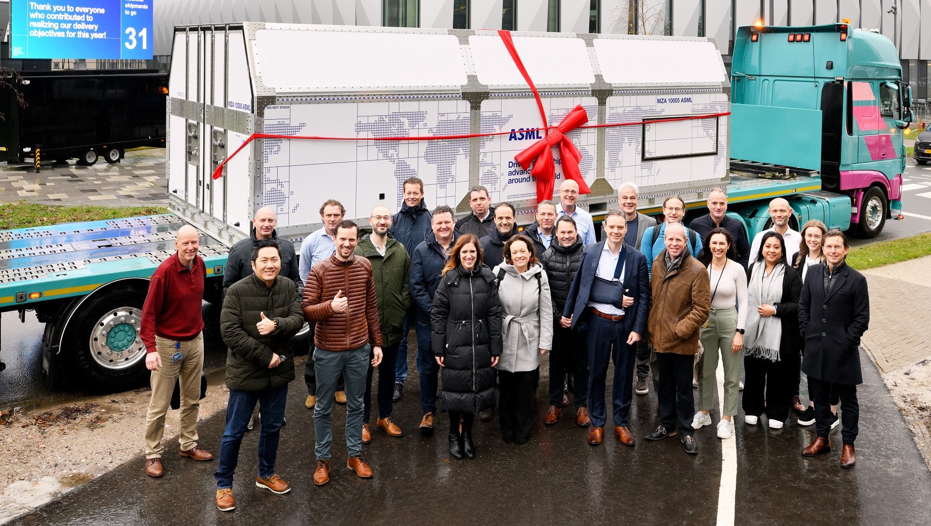 A group of people stand in front of a flatbed truck carrying an ASML shipment box. The box has a red bow tied around it.