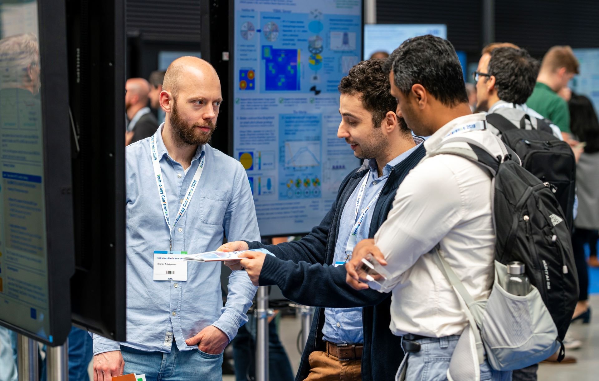 Two men ask another man a question while standing in front of a project poster at the ASML Technology Conference.