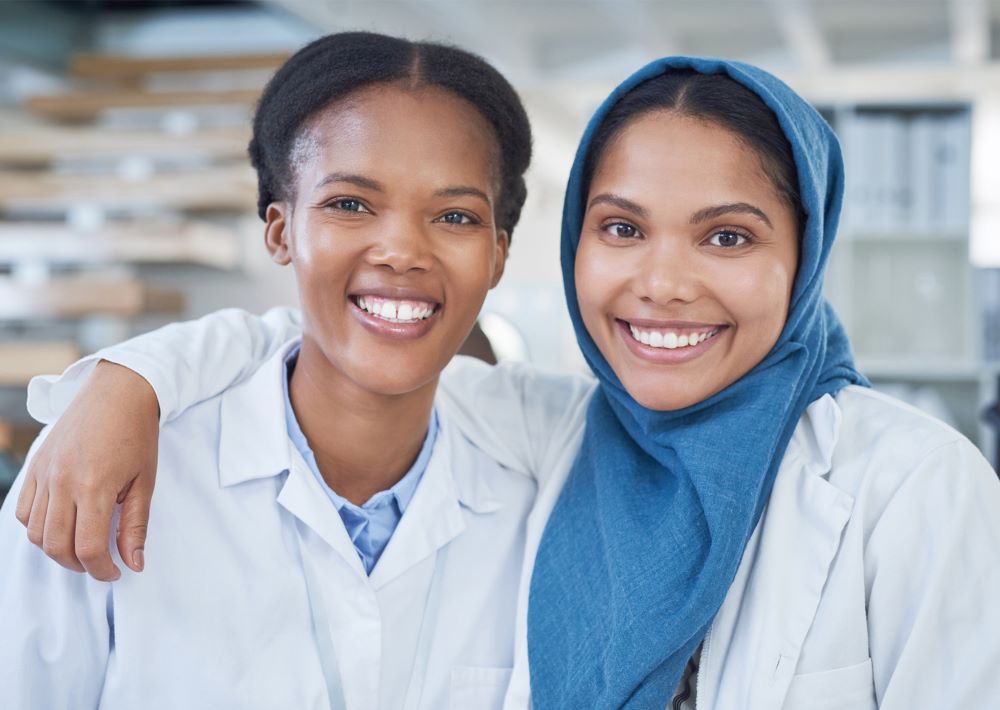 Two female laboratory medicine professionals -- one of whom is Black, and the other of whom wears a hijab -- stand beside each other smiling.
