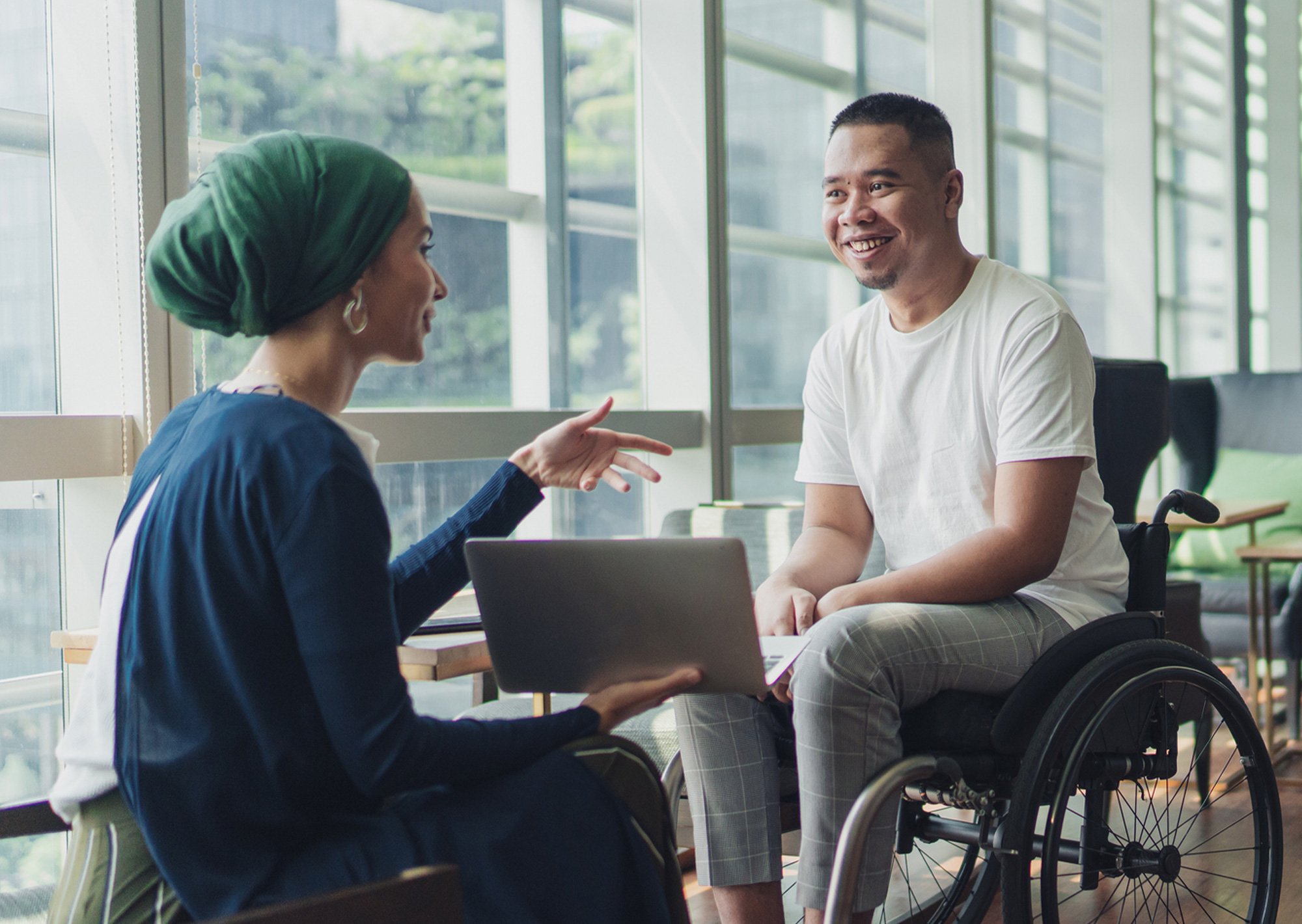 A woman and man in a wheelchair talking beside large windows showing sunlit day
