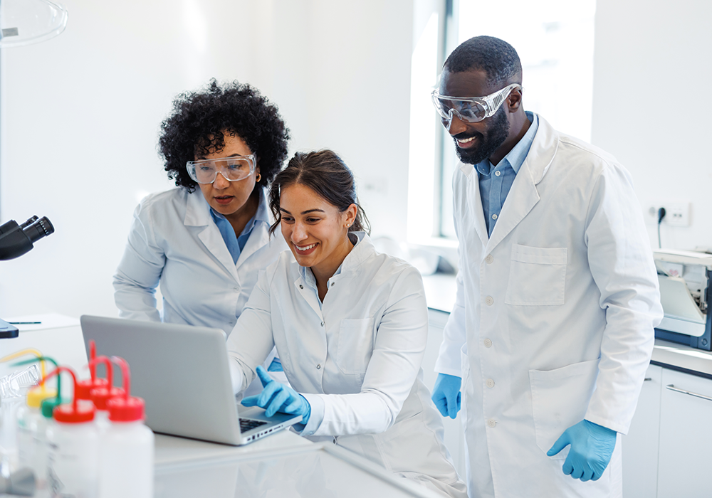Three laboratorians wearing gloves and lab coats look at a laptop.