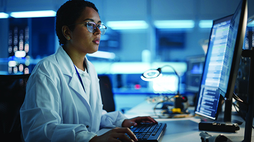 A female laboratorian in a lab, sitting at a desk, and working on a computer