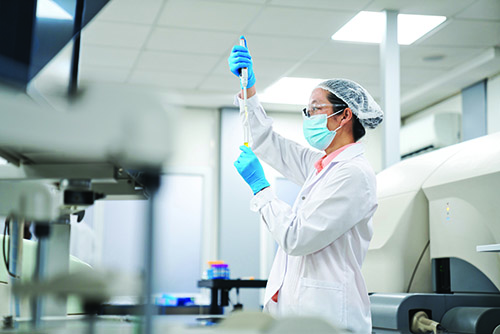 A laboratorian in a hair cap, mask, coat, and gloves, using a pipette to put liquid in a test tube