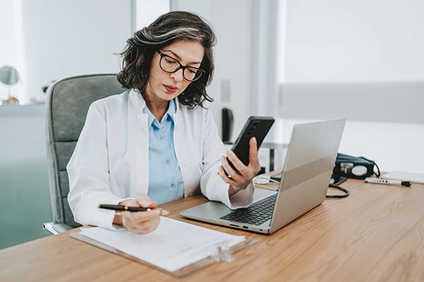 Woman wearing white lab coat works at a desk using a laptop, hand-held device, and paper and pen.