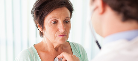 A male physician using a stethoscope and facing a middle-aged woman who looks down places the chestpiece on her chest