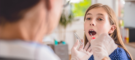 a medical professional with both hands gloved inserts a rapid test swab into a young girl’s mouth