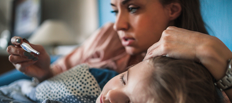 A mother lays beside her child, with one hand on the child’s forehead and with the other holding a thermometer